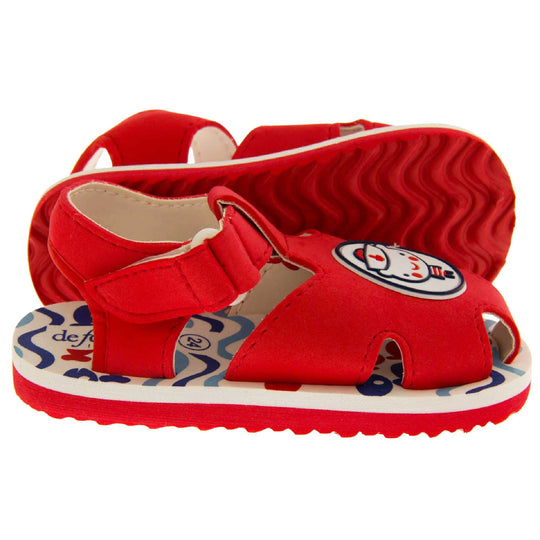 Boys Sandals. Fisherman style sandals with red upper and touch fasten ankle strap. The red upper has a white circle in the centre with a bear dressed as a sailor inside it. The inside of the upper and straps is white, as is the top half of the sole with the foot bed, which has a blue wave pattern on it. The bottom half of the sole is red and has a bumpy grip to the bottom of it.  Both feet from side profile with left foot on its side to show the sole.