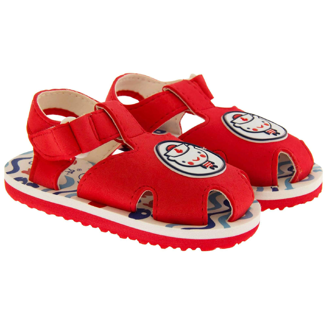 Boys Sandals. Fisherman style sandals with red upper and touch fasten ankle strap. The red upper has a white circle in the centre with a bear dressed as a sailor inside it. The inside of the upper and straps is white, as is the top half of the sole with the foot bed, which has a blue wave pattern on it. The bottom half of the sole is red and has a bumpy grip to the bottom of it. Both shoes next to each other at a slight angle.