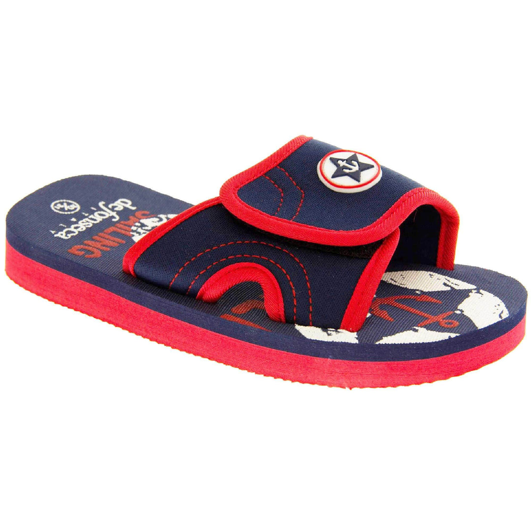 Kids sliders. Slip on slider style sandals. Navy blue touch fasten strap with red around the edge and a white synthetic circle in the centre of the strap with navy anchor in the middle. The foam sole is two toned. The top half is navy, the bottom half is red. Right foot at an angle.