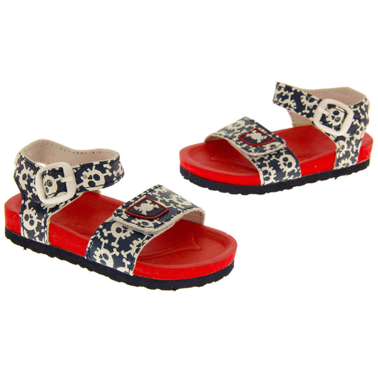 Kids Summer Sandals. Sandals with skull and cross bone pattern on the straps. Strap across the top of the foot by toes and an ankle strap to the back. Both touch fasten with white faux buckle detail on the ankle strap. Top foot bed half of the sole is a red foam, the bottom half is a black firm sole with grip to the bottom. Both shoes in an L shape at a slight angle. 