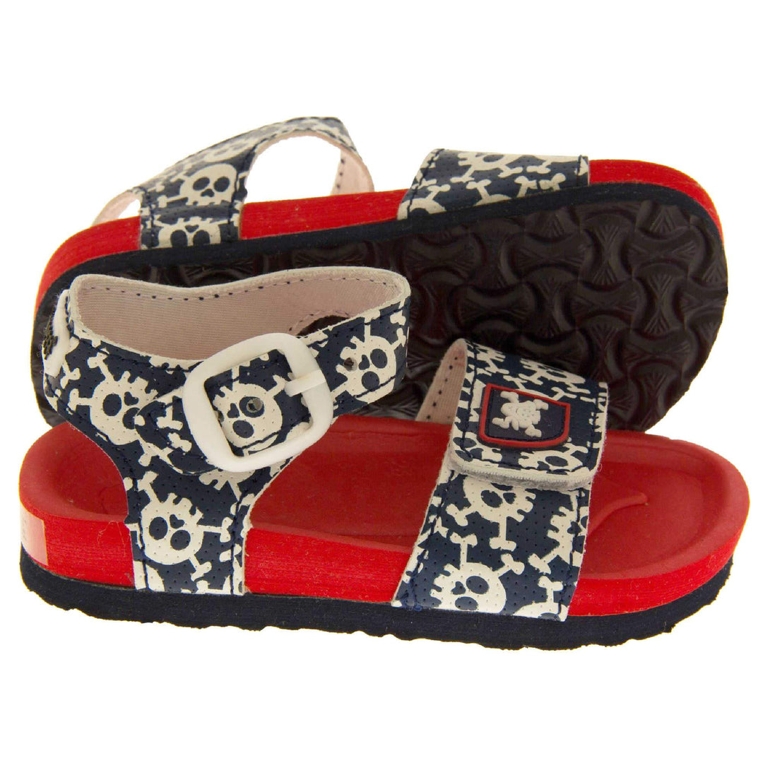 Kids Summer Sandals. Sandals with skull and cross bone pattern on the straps. Strap across the top of the foot by toes and an ankle strap to the back. Both touch fasten with white faux buckle detail on the ankle strap. Top foot bed half of the sole is a red foam, the bottom half is a black firm sole with grip to the bottom. Both feet from side profile with left foot on its side to show the sole.