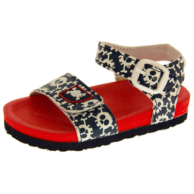 Kids Summer Sandals. Sandals with skull and cross bone pattern on the straps. Strap across the top of the foot by toes and an ankle strap to the back. Both touch fasten with white faux buckle detail on the ankle strap. Top foot bed half of the sole is a red foam, the bottom half is a black firm sole with grip to the bottom. Left foot at an angle