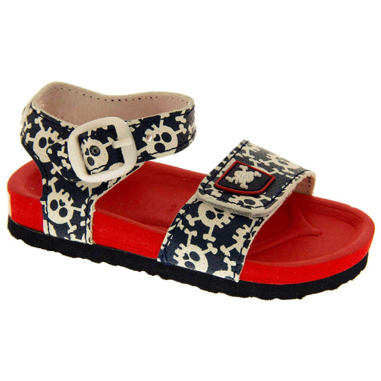 Kids Summer Sandals. Sandals with skull and cross bone pattern on the straps. Strap across the top of the foot by toes and an ankle strap to the back. Both touch fasten with white faux buckle detail on the ankle strap. Top foot bed half of the sole is a red foam, the bottom half is a black firm sole with grip to the bottom. Right foot at an angle