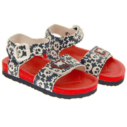 Kids Summer Sandals. Sandals with skull and cross bone pattern on the straps. Strap across the top of the foot by toes and an ankle strap to the back. Both touch fasten with white faux buckle detail on the ankle strap. Top foot bed half of the sole is a red foam, the bottom half is a black firm sole with grip to the bottom. Both shoes next to each other at a slight angle.