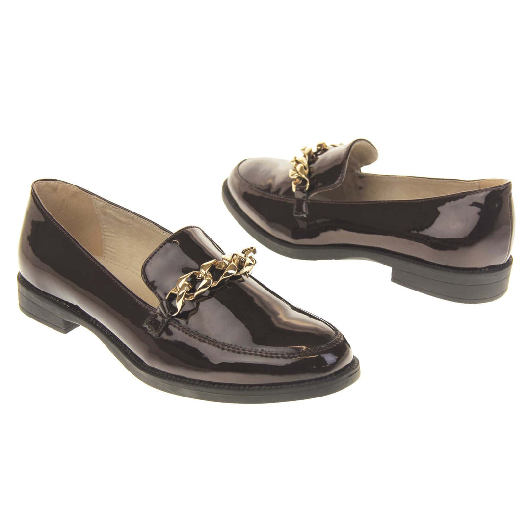 Ladies leather flat shoes. Loafer shoes in a Brogue style with a burgundy faux leather upper. Gold chain detail over the tongue. Real leather lining. Black sole with a slight heel. Both feet at an angle facing top to tail.