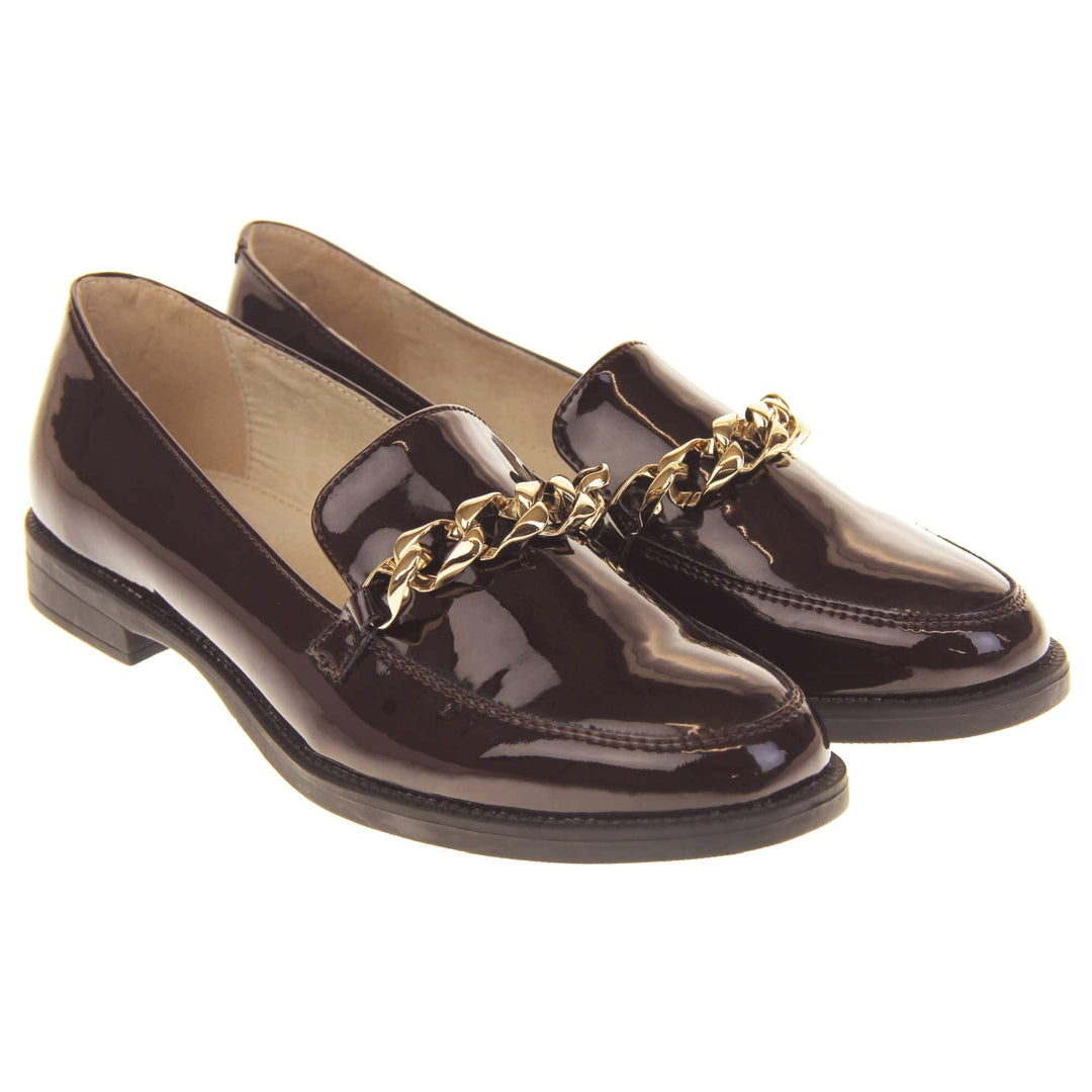 Ladies leather flat shoes. Loafer shoes in a Brogue style with a burgundy faux leather upper. Gold chain detail over the tongue. Real leather lining. Black sole with a slight heel. Both feet together at a slight angle.