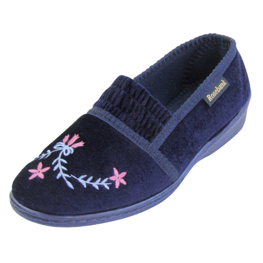 Ladies wedge slippers. Full back slippers in a loafer style. With navy blue velour uppers and a embroidered pale blue and pink flower detail. Ruched velour elasticated gusset. Blue textile lining and piping around the collar. Blue firm sole with a small wedge heel. Left foot at an angle.