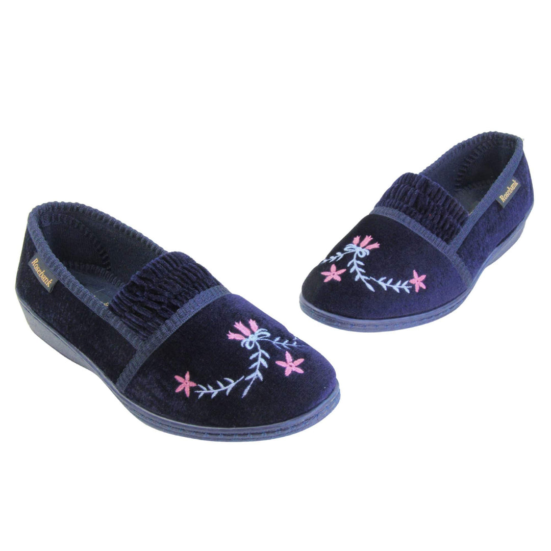 Ladies wedge slippers. Full back slippers in a loafer style. With navy blue velour uppers and a embroidered pale blue and pink flower detail. Ruched velour elasticated gusset. Blue textile lining and piping around the collar. Blue firm sole with a small wedge heel. Both shoes in a V shape with toes almost touching.