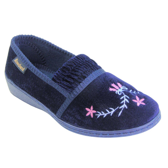 Ladies wedge slippers. Full back slippers in a loafer style. With navy blue velour uppers and a embroidered pale blue and pink flower detail. Ruched velour elasticated gusset. Blue textile lining and piping around the collar. Blue firm sole with a small wedge heel. Right foot at an angle.