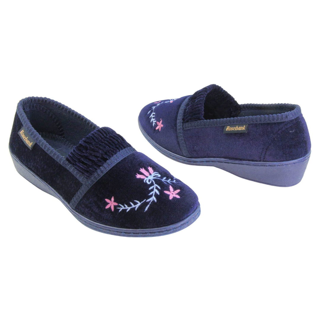 Ladies wedge slippers. Full back slippers in a loafer style. With navy blue velour uppers and a embroidered pale blue and pink flower detail. Ruched velour elasticated gusset. Blue textile lining and piping around the collar. Blue firm sole with a small wedge heel. Both feet at an angle facing top to tail.
