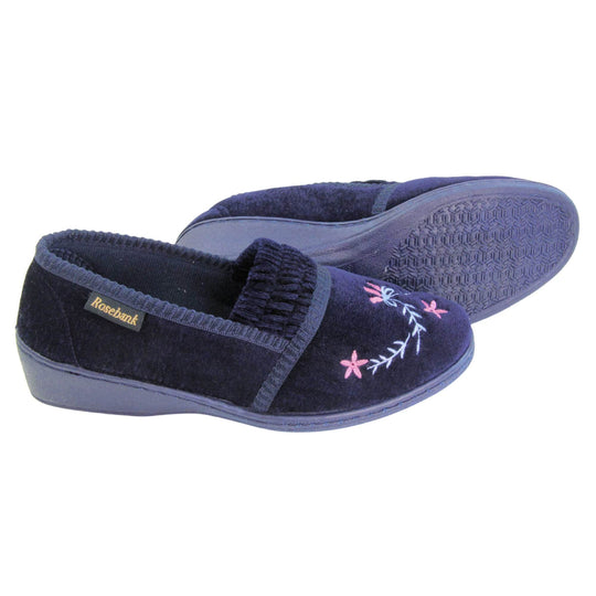 Ladies wedge slippers. Full back slippers in a loafer style. With navy blue velour uppers and a embroidered pale blue and pink flower detail. Ruched velour elasticated gusset. Blue textile lining and piping around the collar. Blue firm sole with a small wedge heel. Both feet from a side profile with the left foot on its side behind the the right foot to show the sole.