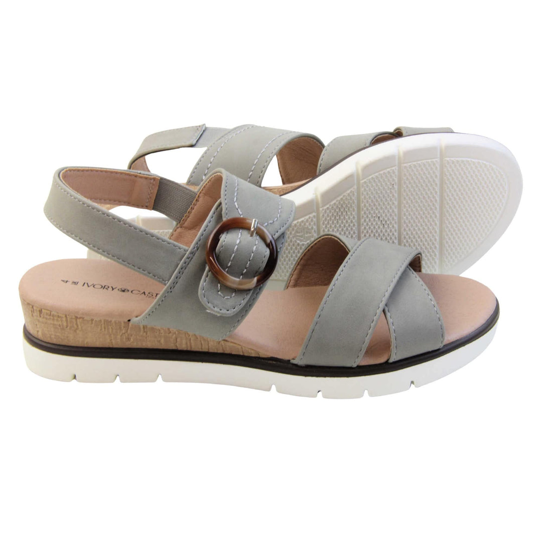 Memory foam wedge sandals. Classic womens strappy sandals with grey textile straps. Dual toe straps that cross over each other. The ankle strap is touch fasten but has a brown buckle detail to look like a buckle fastening. Beige faux leather memory foam insoles. Small wedge heel in cork effect. White outsole with a black rim around the top. Both feet from a side profile with left foot on its side behind the right to show the sole.
