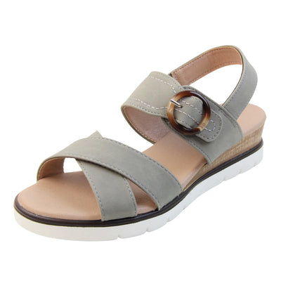 Memory foam wedge sandals. Classic womens strappy sandals with grey textile straps. Dual toe straps that cross over each other. The ankle strap is touch fasten but has a brown buckle detail to look like a buckle fastening. Beige faux leather memory foam insoles. Small wedge heel in cork effect. White outsole with a black rim around the top. Left foot at an angle.