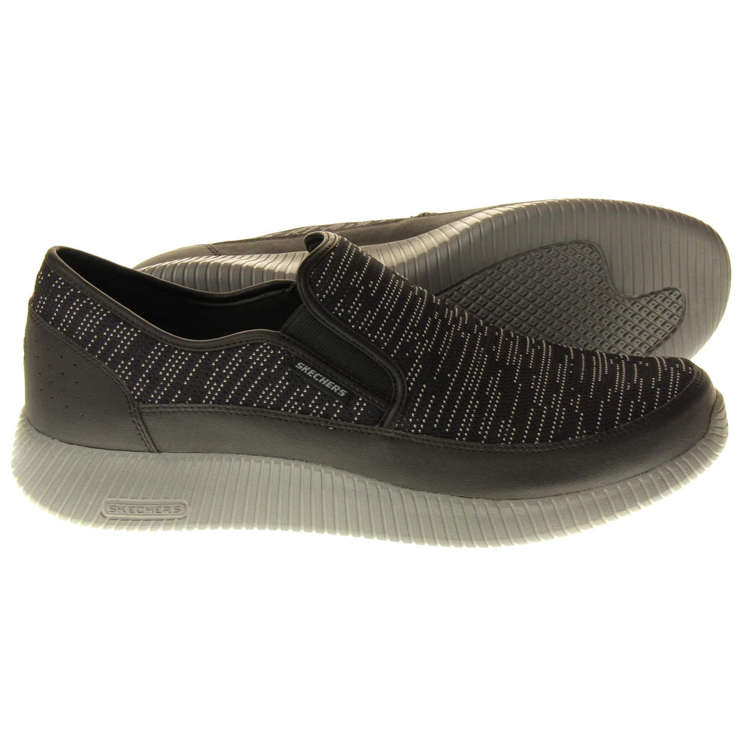 Mens slip on Skechers. Espadrille loafer style shoes with a trainer sole. Black textile upper with white stitch detail. Skechers S logo to the back. Grey thick sole with grip to the bottom. Black textile lining. Both feet from a side profile with the left foot on its side to show the sole.