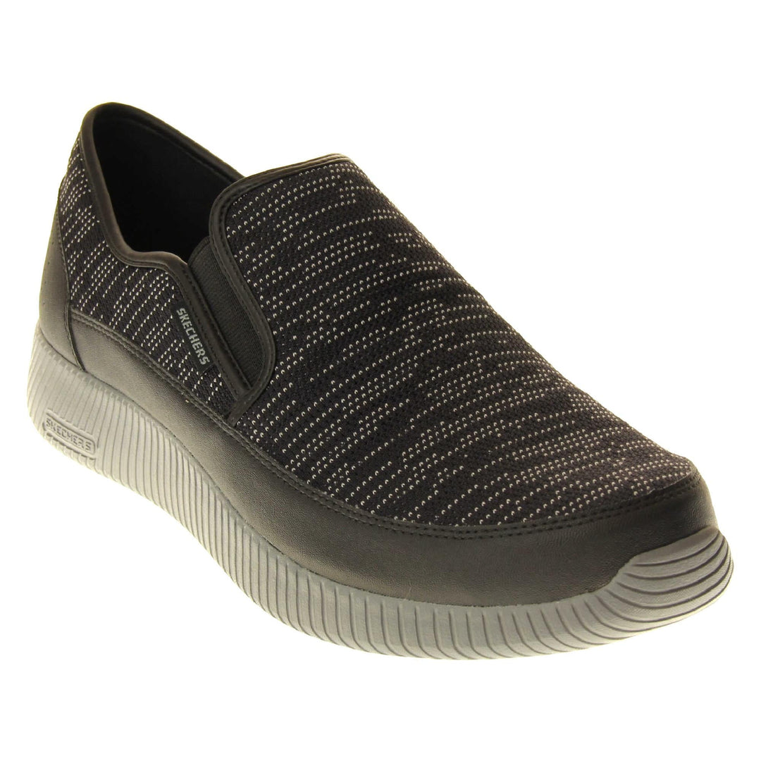 Mens slip on Skechers. Espadrille loafer style shoes with a trainer sole. Black textile upper with white stitch detail. Skechers S logo to the back. Grey thick sole with grip to the bottom. Black textile lining. Right foot at an angle.