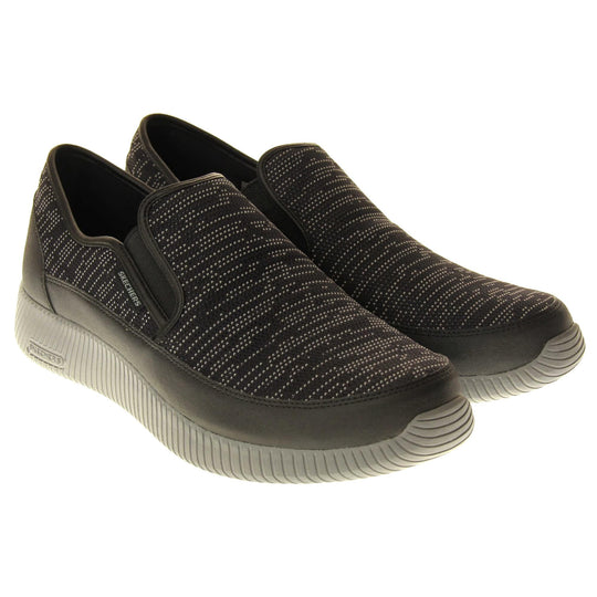 Mens slip on Skechers. Espadrille loafer style shoes with a trainer sole. Black textile upper with white stitch detail. Skechers S logo to the back. Grey thick sole with grip to the bottom. Black textile lining. Both feet together from an angle.