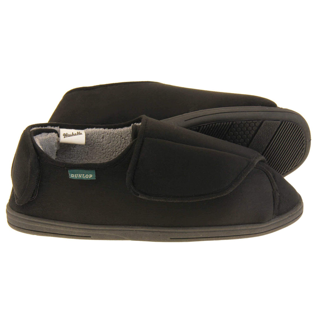 Mens adjustable slippers. Full back slippers with black upper. Adjustable touch fasten strap to the top of the foot and around the back of the heel. Small white label on the outside rim, with Dunlop branding sewn in black. Grey faux fur lining. Firm black sole. Both feet from side profile with left foot on its side to show the sole.