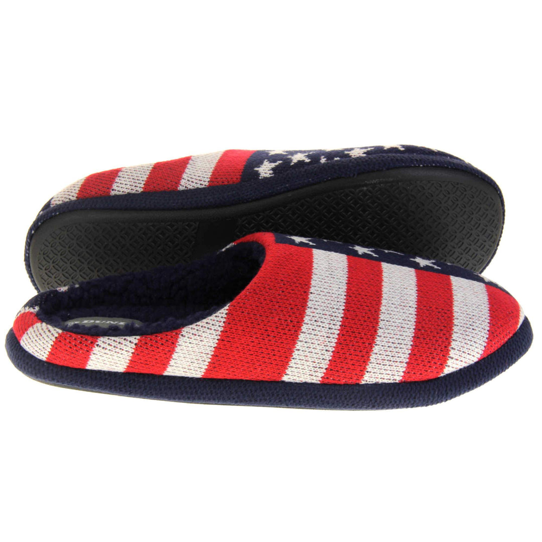 Mens American Flag slippers. Mens slippers in a mule style. With red, white and blue USA flag knit fabric upper. Blue faux fur lining. Black hard synthetic soles with grip to the base. Both feet from a side profile with the left foot on its side to show the sole.