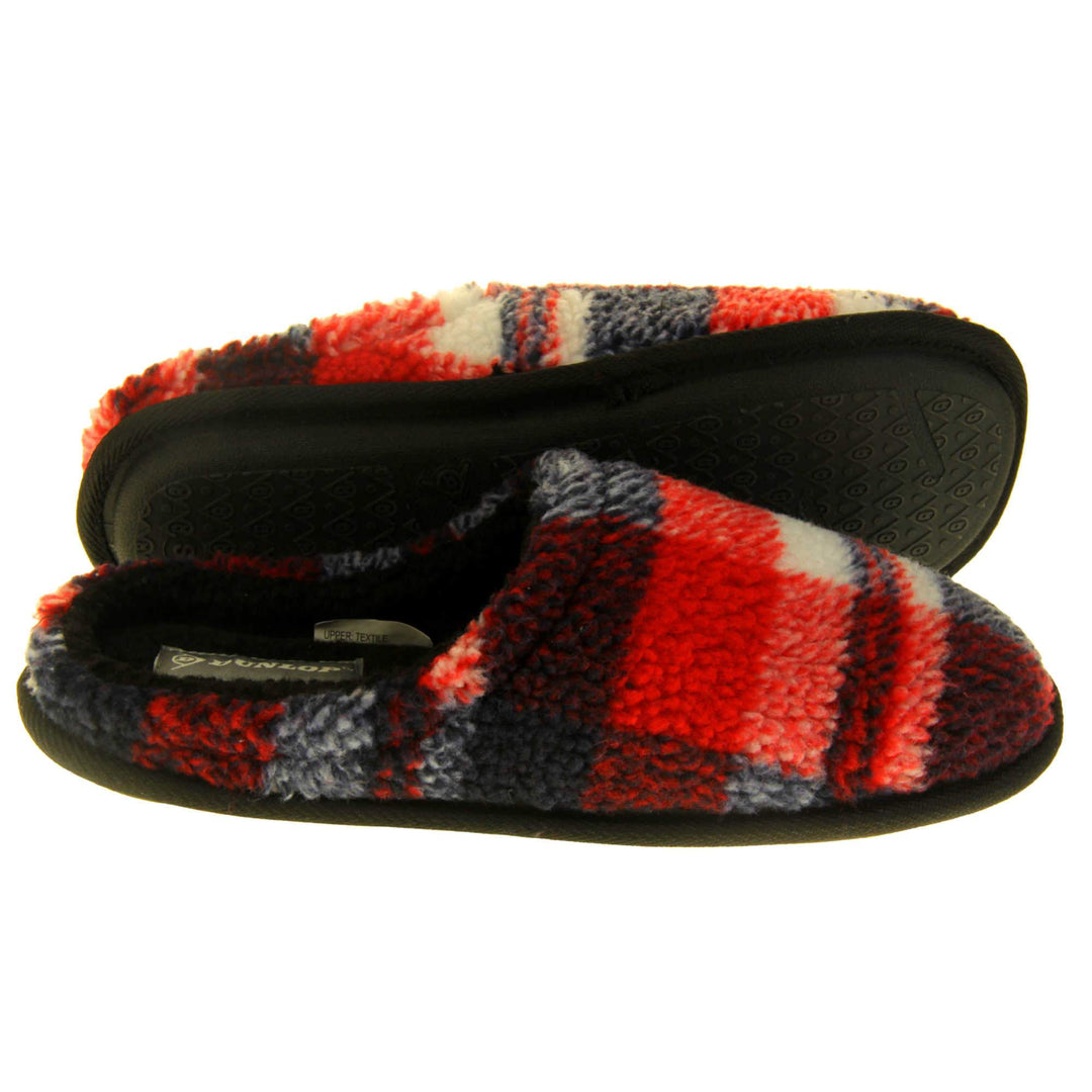 Mens check mule slippers. Mens slippers in a mule style. With red, white and black soft fabric upper. Black fleecy lining. Black hard synthetic soles with grip to the base.  Both feet from a side profile with the left foot on its side to show the sole.