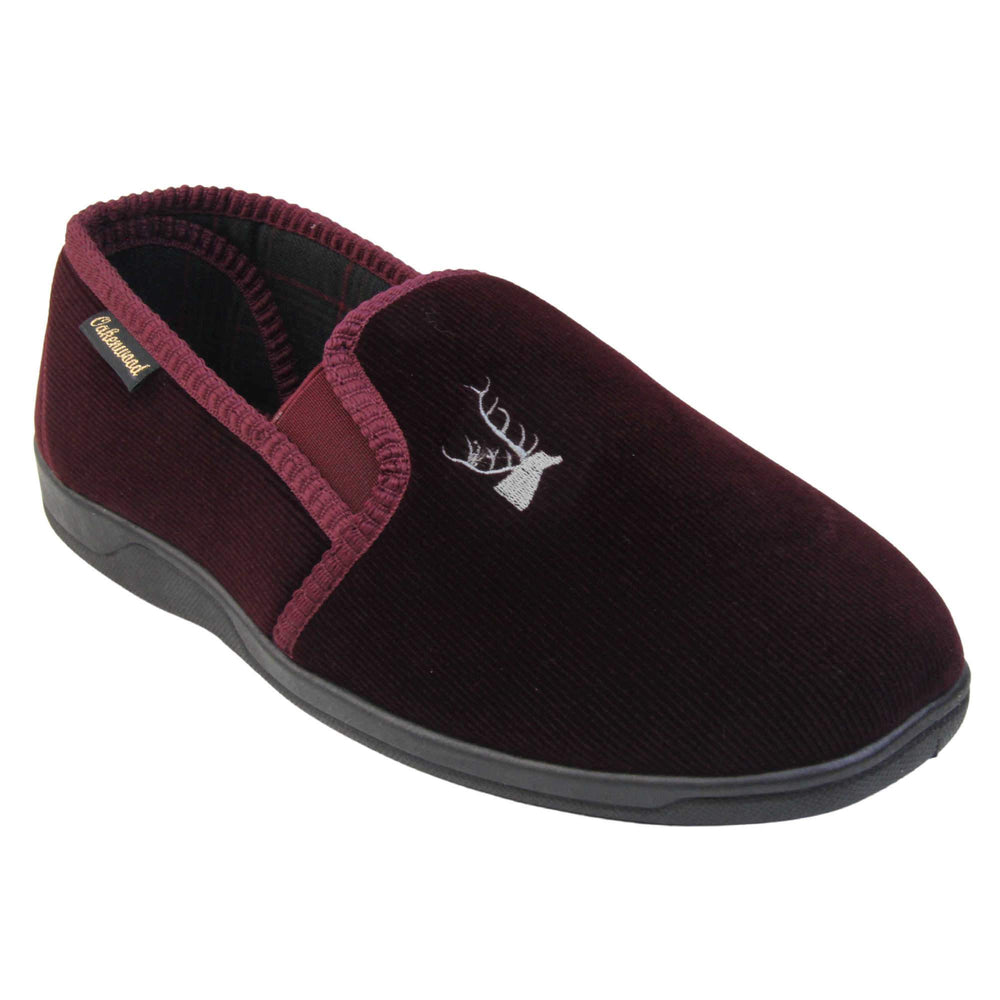 Classic Slippers. Mens full back slippers with a burgundy red velour uppers. Red elasticated panels joining the tongue to the top of the slippers. Grey stag head detail embroidered onto the top of the upper, near the tongue. Small black label on the outside rim, with Oakenwood branding sewn in gold. Plaid textile lining. Right foot at an angle.