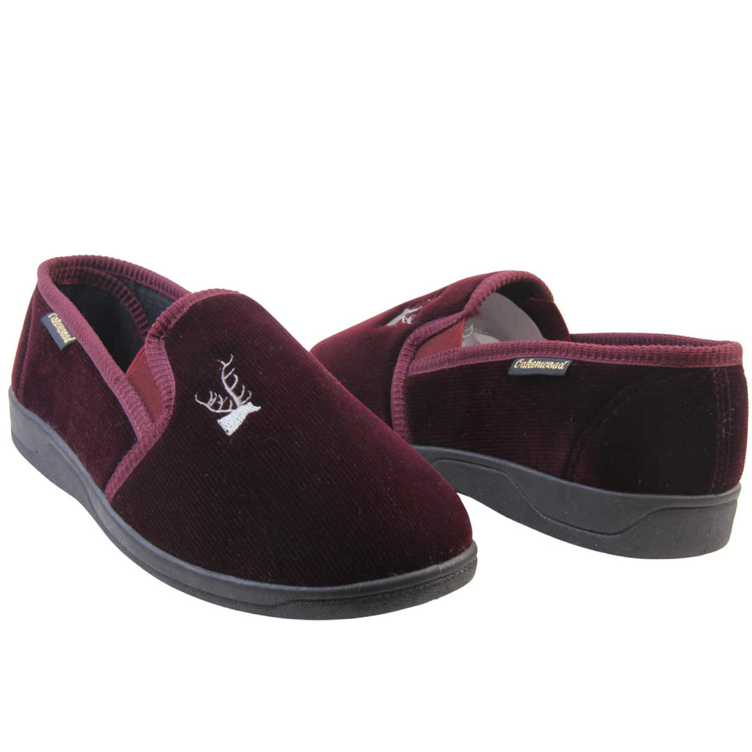 Classic Slippers. Mens full back slippers with a burgundy red velour uppers. Red elasticated panels joining the tongue to the top of the slippers. Grey stag head detail embroidered onto the top of the upper, near the tongue. Small black label on the outside rim, with Oakenwood branding sewn in gold. Plaid textile lining. Both feet facing top to tail, at an angle.