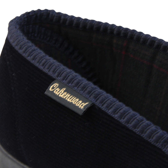 Mens Comfortable Slippers. Full back slippers for men with a navy blue velour uppers. Navy elasticated panels joining the tongue to the top of the slippers. Grey stag head detail embroidered onto the top of the upper, near the tongue. Small navy label on the outside rim, with Oakenwood branding sewn in gold. Navy textile lining. Close up of the back part of the foot to show the label with branding and the lining of the slippers.