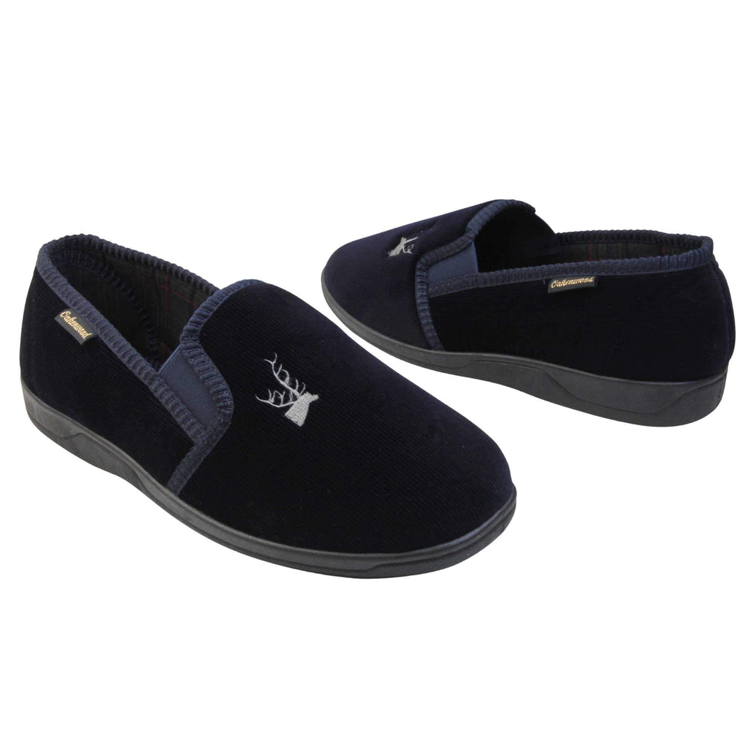 Mens Comfortable Slippers. Full back slippers for men with a navy blue velour uppers. Navy elasticated panels joining the tongue to the top of the slippers. Grey stag head detail embroidered onto the top of the upper, near the tongue. Small navy label on the outside rim, with Oakenwood branding sewn in gold. Navy textile lining. Both feet facing top to tail, at an angle.