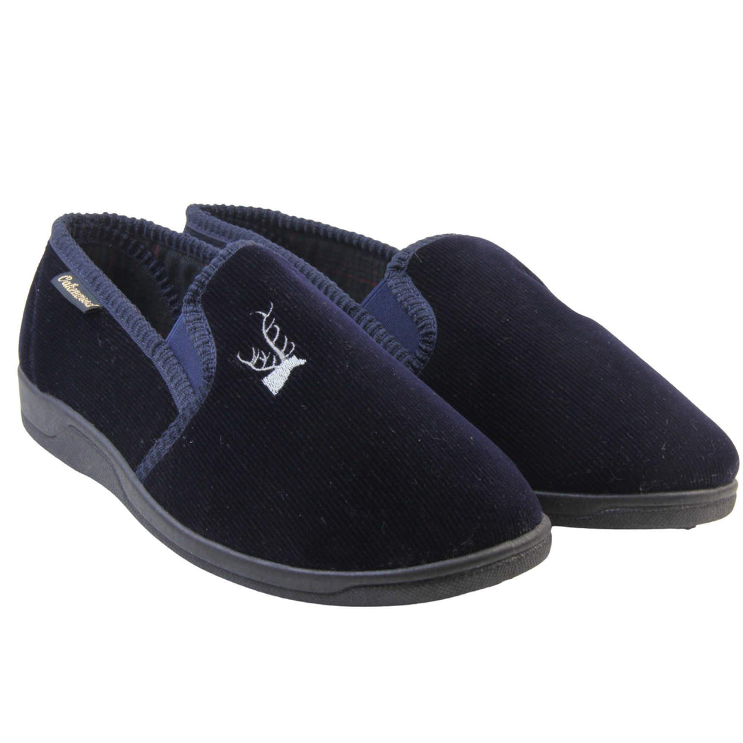 Mens Comfortable Slippers. Full back slippers for men with a navy blue velour uppers. Navy elasticated panels joining the tongue to the top of the slippers. Grey stag head detail embroidered onto the top of the upper, near the tongue. Small navy label on the outside rim, with Oakenwood branding sewn in gold. Navy textile lining. Both feet together at an angle.