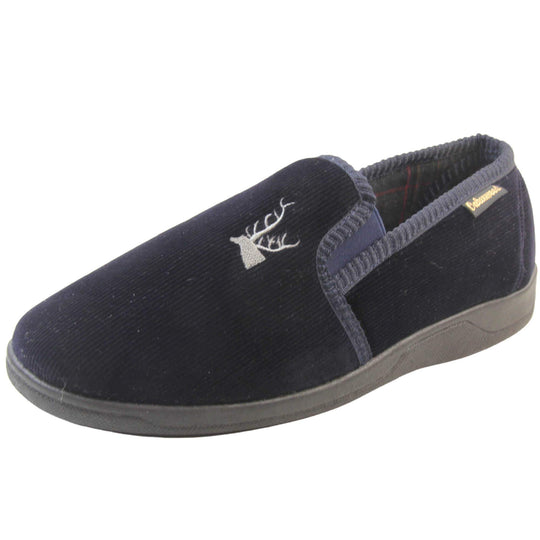 Mens Comfortable Slippers. Full back slippers for men with a navy blue velour uppers. Navy elasticated panels joining the tongue to the top of the slippers. Grey stag head detail embroidered onto the top of the upper, near the tongue. Small navy label on the outside rim, with Oakenwood branding sewn in gold. Navy textile lining. Left foot at an angle.