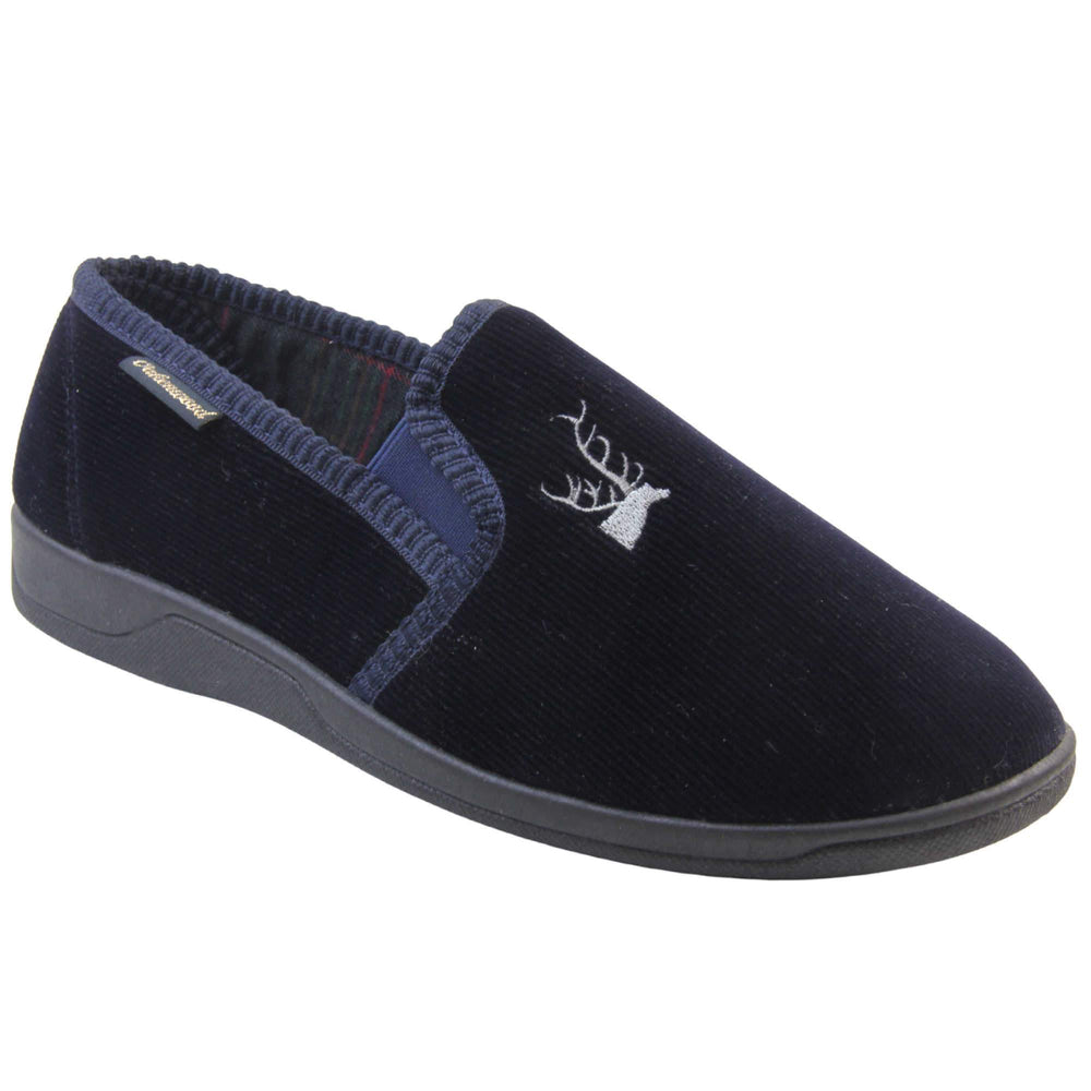 Mens Comfortable Slippers. Full back slippers for men with a navy blue velour uppers. Navy elasticated panels joining the tongue to the top of the slippers. Grey stag head detail embroidered onto the top of the upper, near the tongue. Small navy label on the outside rim, with Oakenwood branding sewn in gold. Navy textile lining. Right foot at an angle