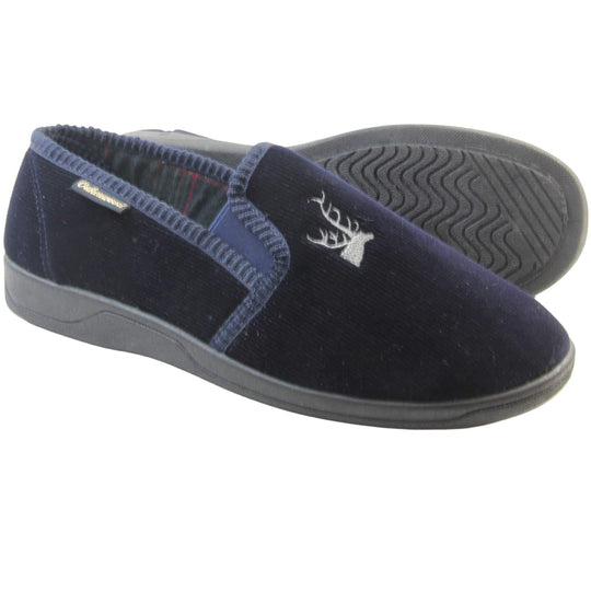 Mens Comfortable Slippers. Full back slippers for men with a navy blue velour uppers. Navy elasticated panels joining the tongue to the top of the slippers. Grey stag head detail embroidered onto the top of the upper, near the tongue. Small navy label on the outside rim, with Oakenwood branding sewn in gold. Navy textile lining. Both feet from side profile with left foot on its side to show the sole.