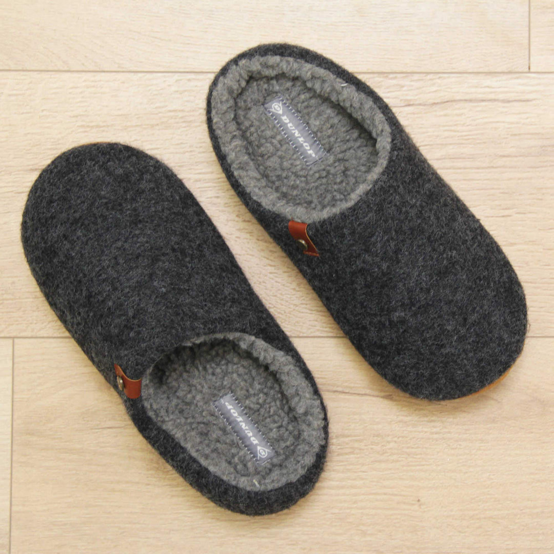 Mens memory foam slippers. Mule style slippers with a black felt upper with brown rim around the base and a brown faux leather strap with stud detail along the collar of the slipper. Grey Wool effect faux fur lining with a grey Dunlop label on the middle of the insole. Firm black sole with wavy lines for grip. Lifestyle photo from above of both slippers top to tale on a wood floor.