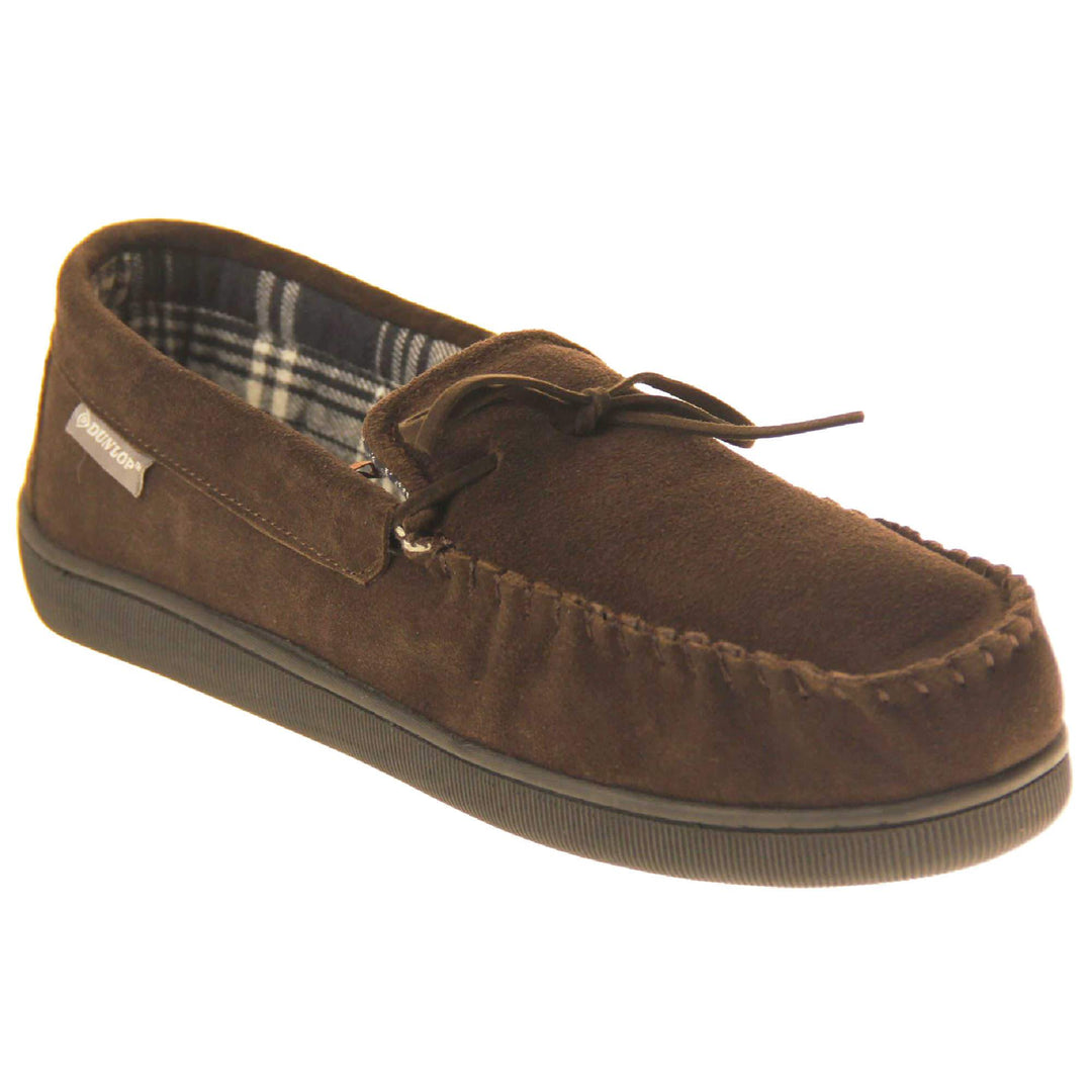 Mens moccasin slippers. Moccasin style slipper with brown suede upper and leather bow to the top. Grey Dunlop label to the outside. Grey and white plaid textile lining. Black rubber sole. Right foot at an angle.