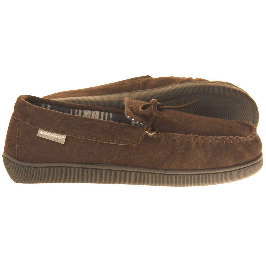 Mens moccasin slippers. Moccasin style slipper with brown suede upper and leather bow to the top. Grey Dunlop label to the outside. Grey and white plaid textile lining. Black rubber sole. Both feet from a side profile with the left foot on its side to show the sole.