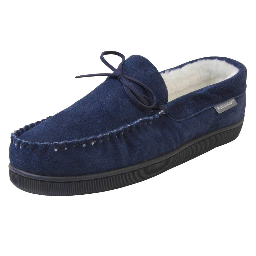 Mens moccasin slippers. Closed back slippers in a moccasin style with navy blue suede leather upper and bow. Cream faux fur lining. Thick black sole. Left foot at an angle.