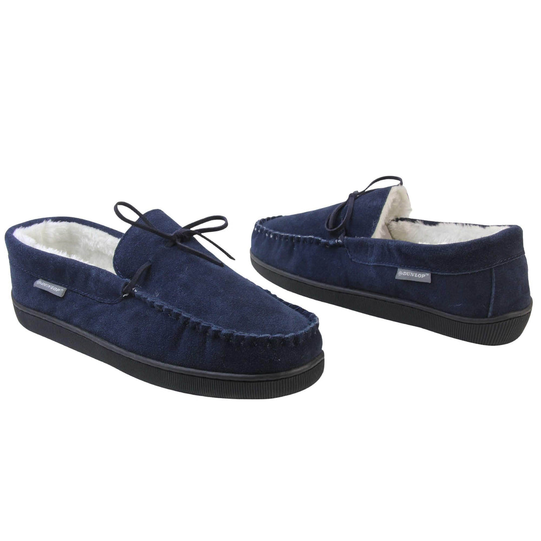 Mens moccasin slippers. Closed back slippers in a moccasin style with navy blue suede leather upper and bow. Cream faux fur lining. Thick black sole. Both shoes at an angle spaced about an inch apart, facing top to tail at an angle.