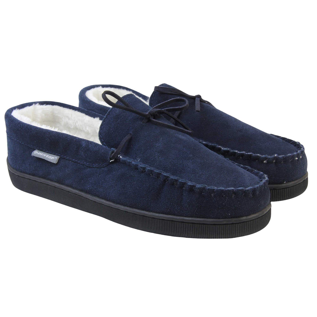 Mens moccasin slippers. Closed back slippers in a moccasin style with navy blue suede leather upper and bow. Cream faux fur lining. Thick black sole. Both feet together at an angle.
