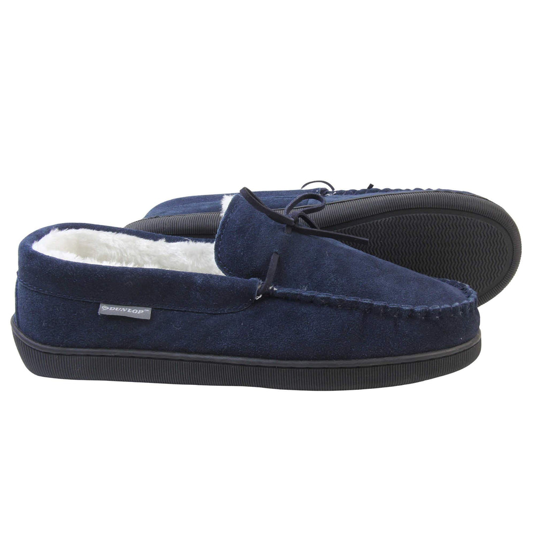 Mens moccasin slippers. Closed back slippers in a moccasin style with navy blue suede leather upper and bow. Cream faux fur lining. Thick black sole. Both feet from a side profile with the left foot on its side to show the sole.