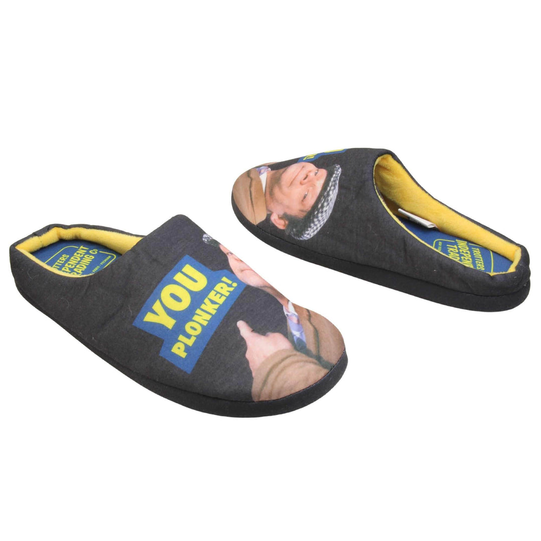 Mens mule novelty slippers. Mens slippers in a mule style. With black fabric upper with Del Boy from Only Fools and Horses printed on it and pointing. The words you plonker! in bold yellow font outlined in blue above his pointing finger. Blue fabric insole with yellow Totters independent trading logo on. Yellow fleece lining. Black hard synthetic soles with bumpy grip to the base. Both feet from an angle facing top to tail.