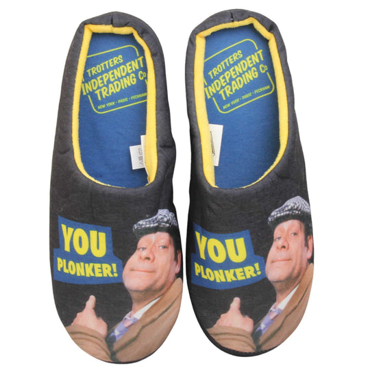 Mens mule novelty slippers. Mens slippers in a mule style. With black fabric upper with Del Boy from Only Fools and Horses printed on it and pointing. The words you plonker! in bold yellow font outlined in blue above his pointing finger. Blue fabric insole with yellow Totters independent trading logo on. Yellow fleece lining. Black hard synthetic soles with bumpy grip to the base. Both feet together from above.