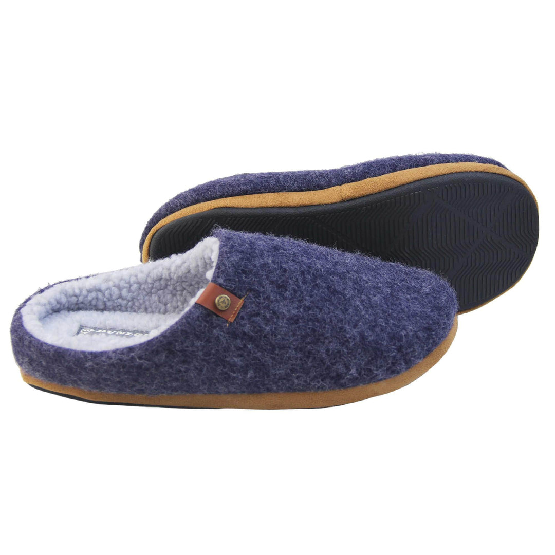 Mens mule slippers. Mule style slippers with a navy blue felt upper with brown rim around the base and a brown faux leather strap with stud detail along the collar of the slipper. Grey Wool effect faux fur lining with a grey Dunlop label on the middle of the insole. Firm black sole with wavy lines for grip. Both feet from a side profile with left foot on its side to show the sole.