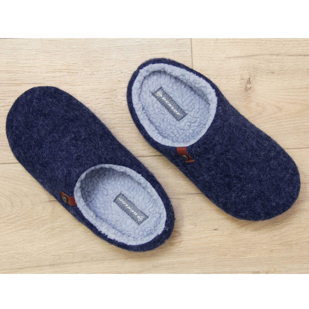Mens mule slippers. Mule style slippers with a navy blue felt upper with brown rim around the base and a brown faux leather strap with stud detail along the collar of the slipper. Grey Wool effect faux fur lining with a grey Dunlop label on the middle of the insole. Firm black sole with wavy lines for grip. Lifestyle photo from above of both slippers top to tale on a wood floor.