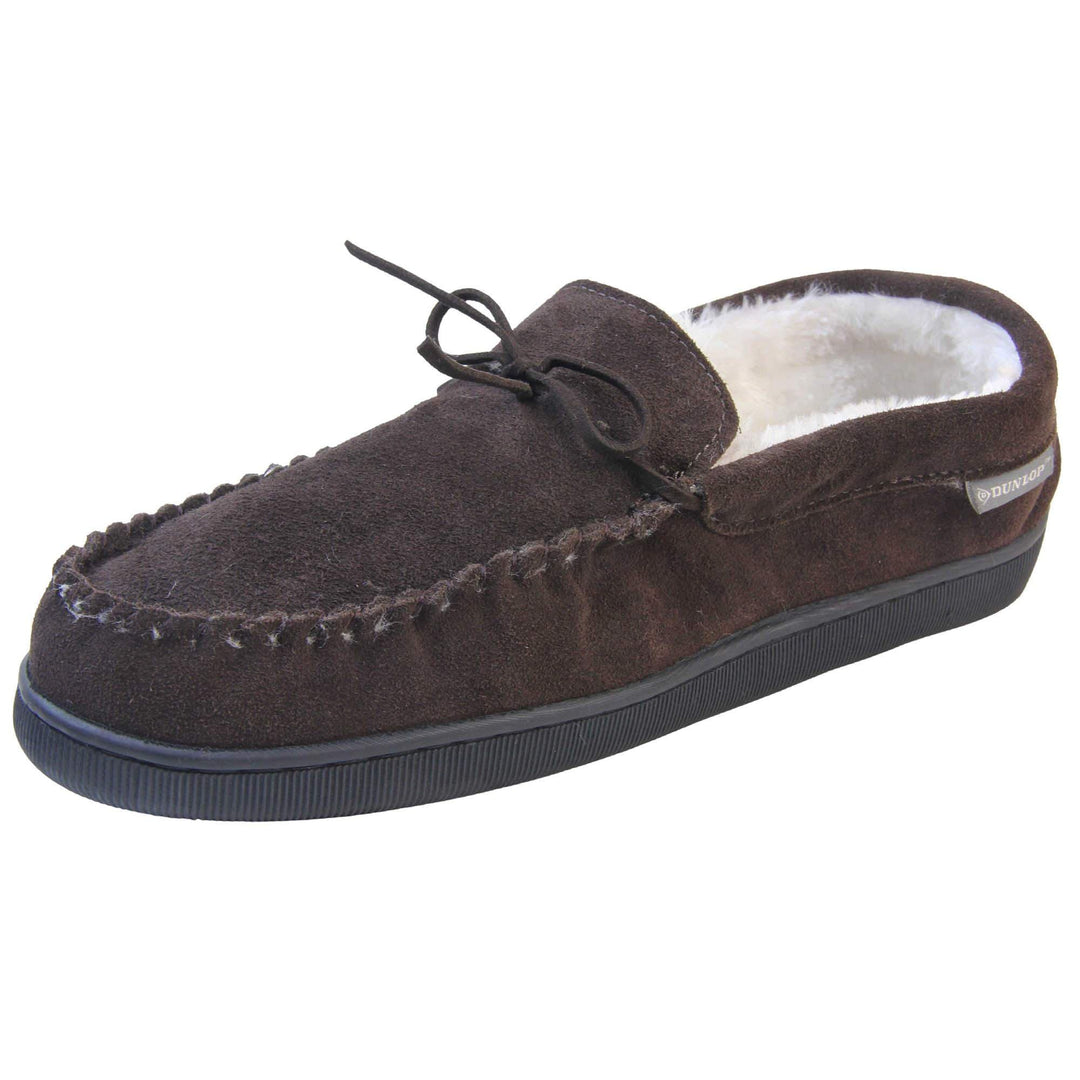 Mens suede moccasin slippers. Closed back slippers in a moccasin style with dark brown suede leather upper and bow. Cream faux fur lining. Thick black sole. Left foot at an angle.