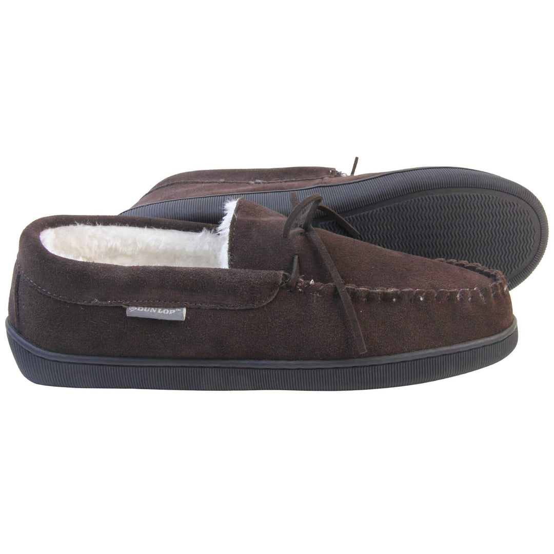 Mens suede moccasin slippers. Closed back slippers in a moccasin style with dark brown suede leather upper and bow. Cream faux fur lining. Thick black sole. Both feet from a side profile with the left foot on its side to show the sole.