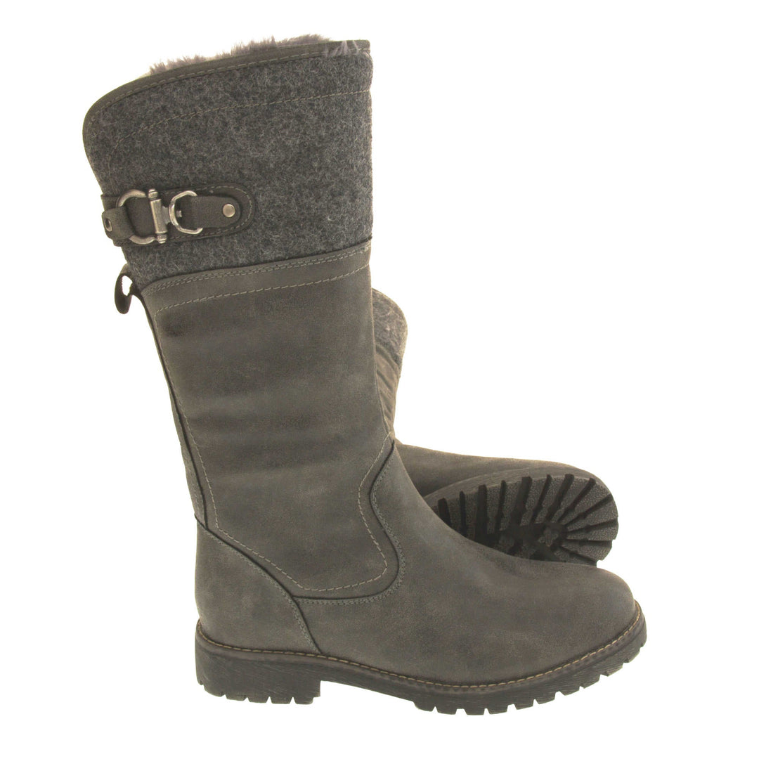 Mid calf winter boots. Women's boot in a below the knee style. With a grey faux leather upper and grey felt effect collar with grey buckle detail. Grey sole with chunky grip to the bottom. Zip fastening to the inside leg. Grey faux fur lining. Both feet from a side profile with the left foot on its side to show the sole.