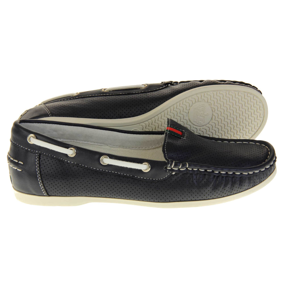 Navy deck shoes. Women's boat shoes with a navy upper and tiny dot cut-outs. White lace detail running around the outside of the collar. White stitching detailing around the top of the shoe. Cream leather lining and sole. Both feet from a side profile with the left foot on its side behind the the right foot to show the sole.