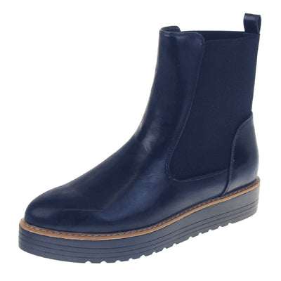 Navy flat ankle boots. Faux leather, tall Chelsea boot style with a navy blue coloured upper. Blue elasticated panels at the ankles and a navy loop at the heel to help pull them on. Black coloured low platform sole. Left foot at an angle