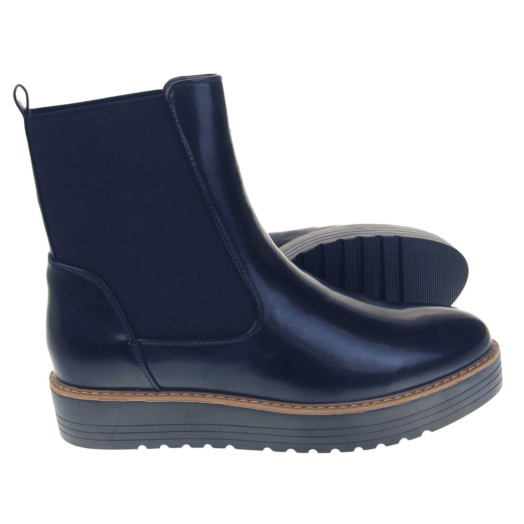 Navy flat ankle boots. Faux leather, tall Chelsea boot style with a navy blue coloured upper. Blue elasticated panels at the ankles and a navy loop at the heel to help pull them on. Black coloured low platform sole. Both feet from a side profile with the left foot on its side to show the sole.