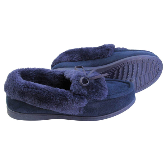 Navy moccasin slippers. Moccasin style slipper with a navy blue faux suede upper and bow to the top. Navy faux fur collar, tongue and lining. Navy rubber sole. Both feet from a side profile with the left foot on its side behind the the right foot to show the sole.