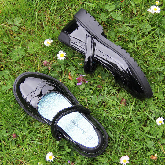 Older girls school shoes. Mary Jane style shoes with black patent faux leather uppers. With a touch fasten strap over the foot and brogue detailing around the top of the shoe. Black textile lining with a metallic blue insole. Chunky black sole with slight heel. Lifestyle photo with both shoes on grass from above facing top to tail with one on its side.